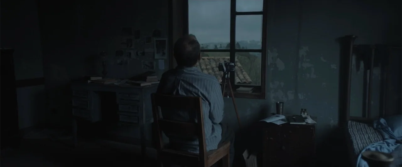 My Neighbor Adolf (2022) movie still: medium — A dark, sparsely furnished room contains a desk and a bed in shadow. A man sits on a wooden chair…