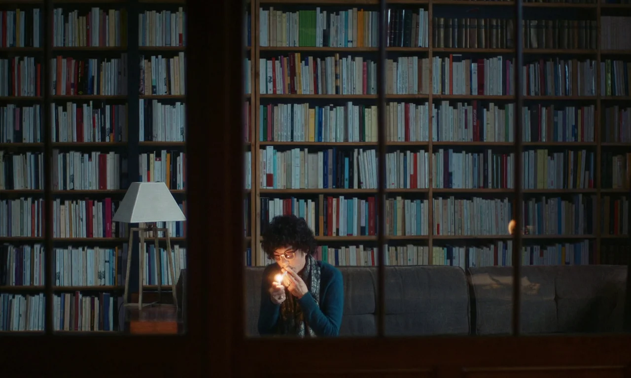 Little Girl Blue (2023) movie still: wide — A person sits on a sofa in front of a wall of filled bookshelves. The person lights a cigarette with a…