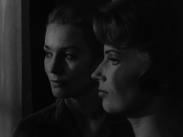The Silence (1963) movie still: closeup — Two women stand in close proximity in a dark, shadow-filled interior space. They gaze ahead with neutral…