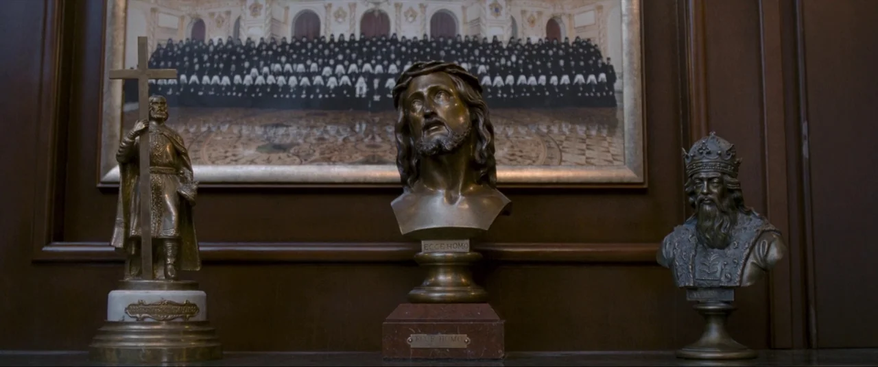 Leviathan (2014) movie still: medium — Three bronze sculptures stand on a wooden surface before a framed photograph of clergy. The sculptures depict…