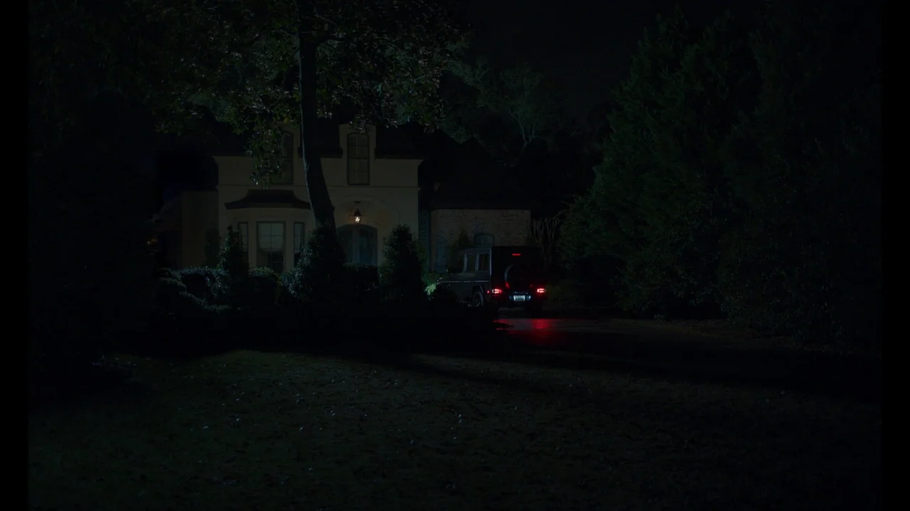 The Card Counter (2021) movie still: establishing — A dark, two-story house stands at the end of a driveway surrounded by trees. A black SUV is…
