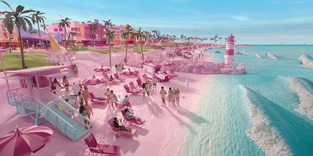 Barbie (2023) movie still: wide — A vibrant pink sand beach features a striped lighthouse and stylized pastel architecture. Small groups of people…