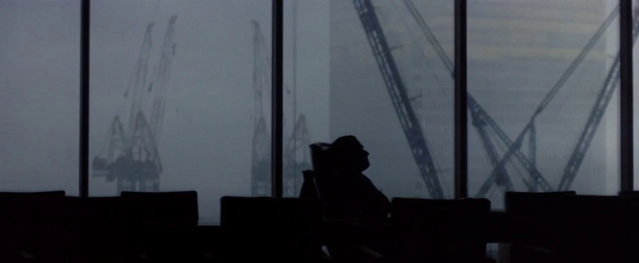 Klute (1971) movie still: wide — A high-rise conference room overlooks a rainy urban skyline with multiple construction cranes visible. A man sits…