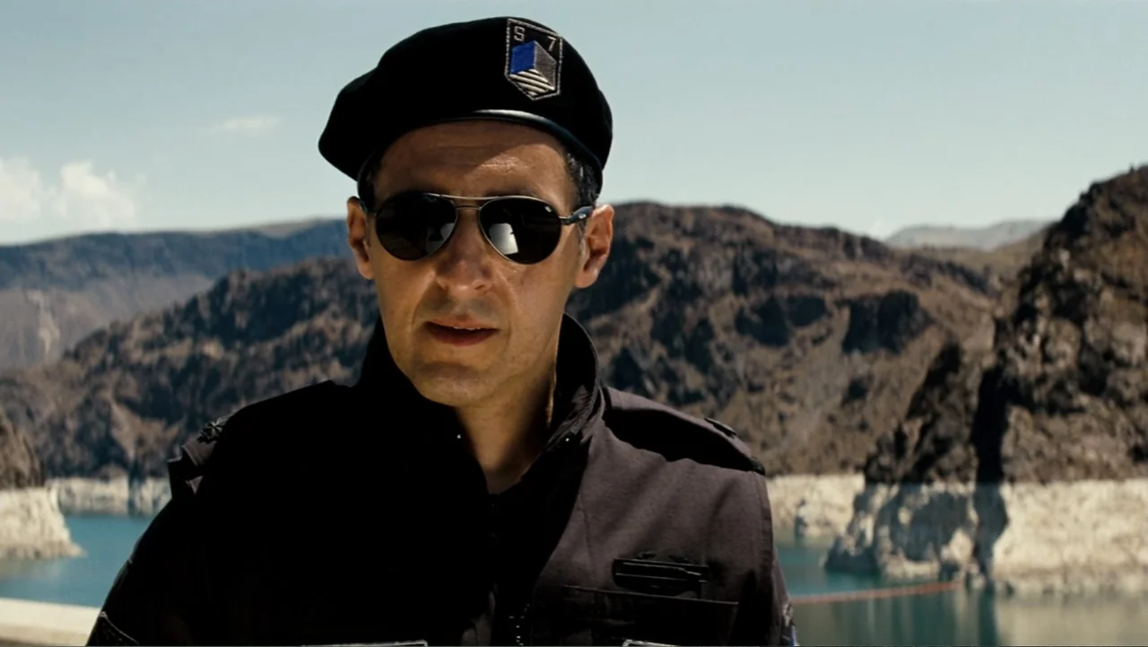 Transformers (2007) movie still: medium — A man in a black tactical uniform stands in front of a water reservoir. The background shows a concrete…