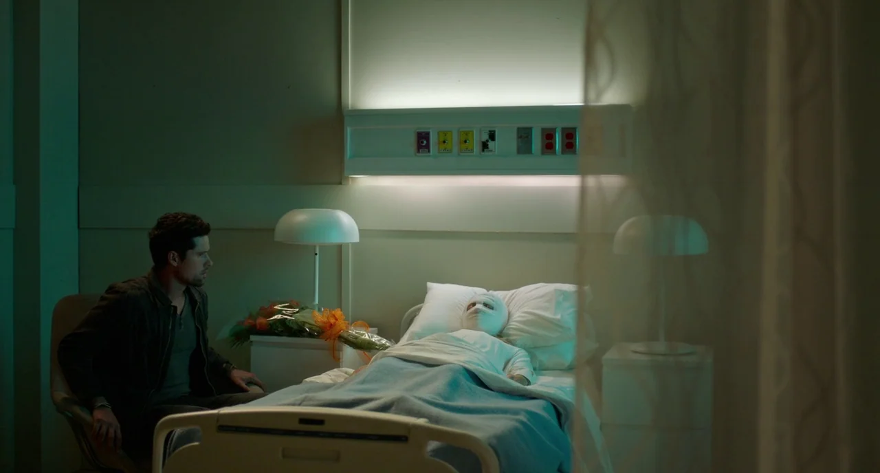 Rabid (2019) movie still: medium — A quiet hospital room features a bed occupied by a person with their head bandaged. A man sits beside the patient…