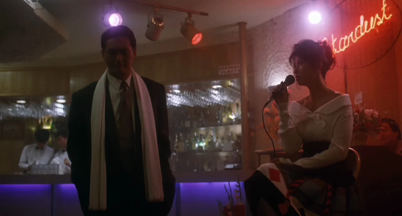 The Killer (1989) movie still: medium — A dimly lit lounge with a neon Stardust sign on the wall behind the stage. A man stands while a woman…