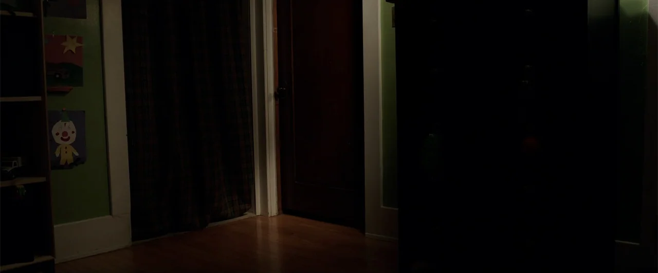 Lights Out (2016) movie still: establishing — A dimly lit room features a dark wood door and green walls with drawings. The floor is covered by dark…