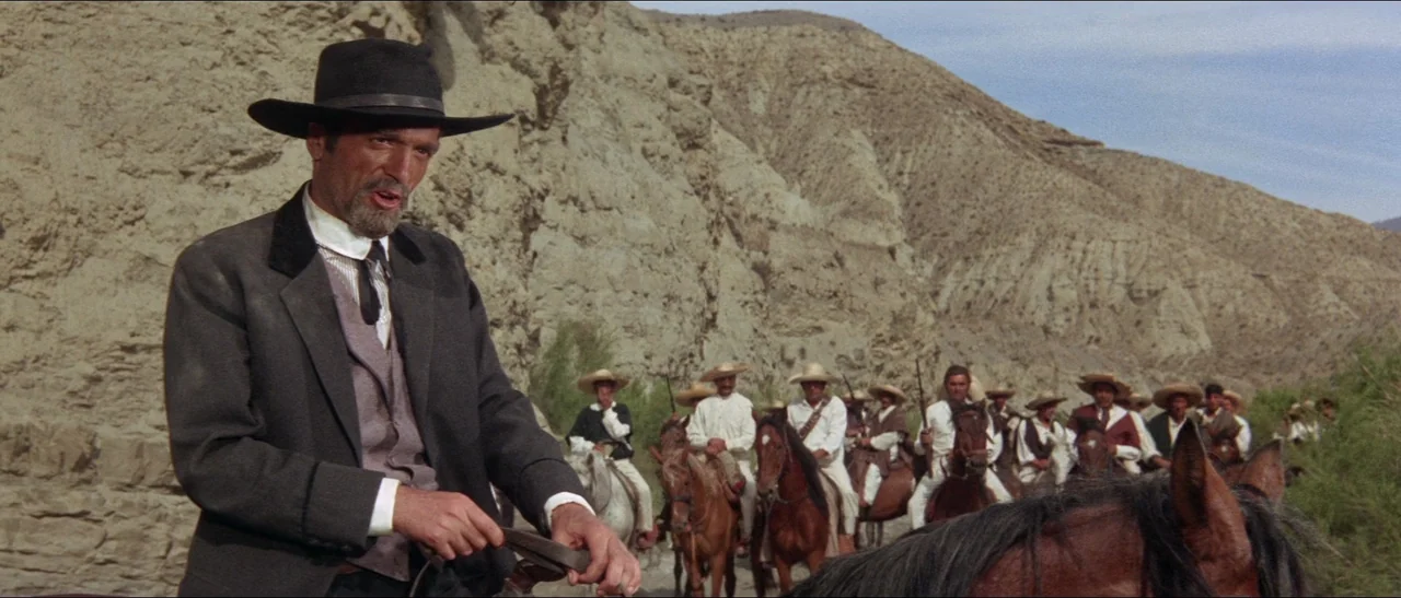 Texas, Adios (1966) movie still: medium — A group of armed men on horseback traverse a dry, rocky desert canyon landscape. The lead rider in a suit…