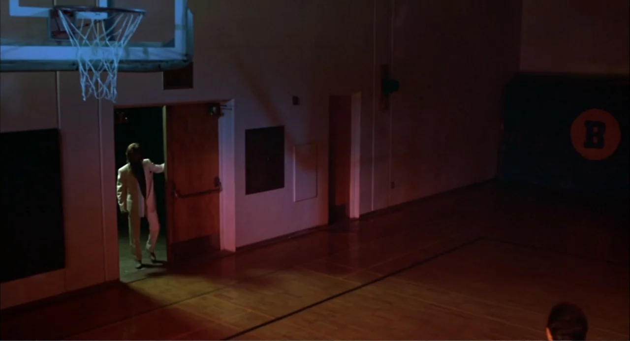Teen Wolf (1985) movie still: wide — A dark gymnasium with polished wooden floors and a blue padded wall with a logo. A man in a light-colored suit…
