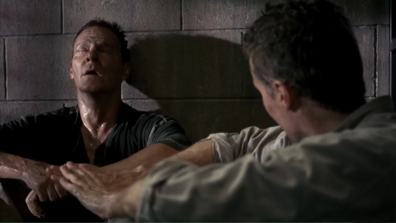 Behind Enemy Lines (2001) movie still: medium — A spartan room is lined with grey cinder block walls and dim lighting. One man rests against the…