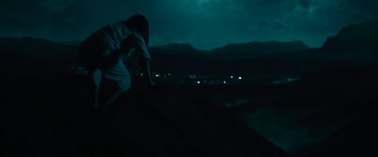 Wildling (2018) movie still: wide — A dark rocky hilltop overlooks a valley with distant glowing lights at night. A person in white clothing…