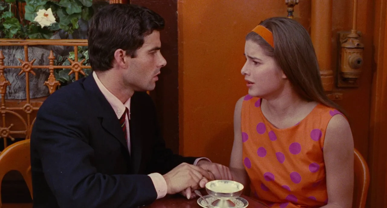 The Umbrellas Of Cherbourg (1964) movie still: medium — A small table sits before a plain wall decorated with a hanging plant. A man and woman sit…