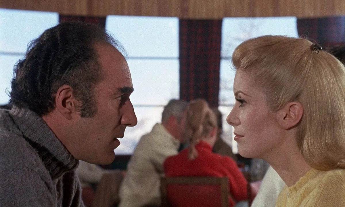 Belle De Jour (1967) movie still: medium — A bright indoor restaurant setting features large windows and patterned curtains in the background. A man…