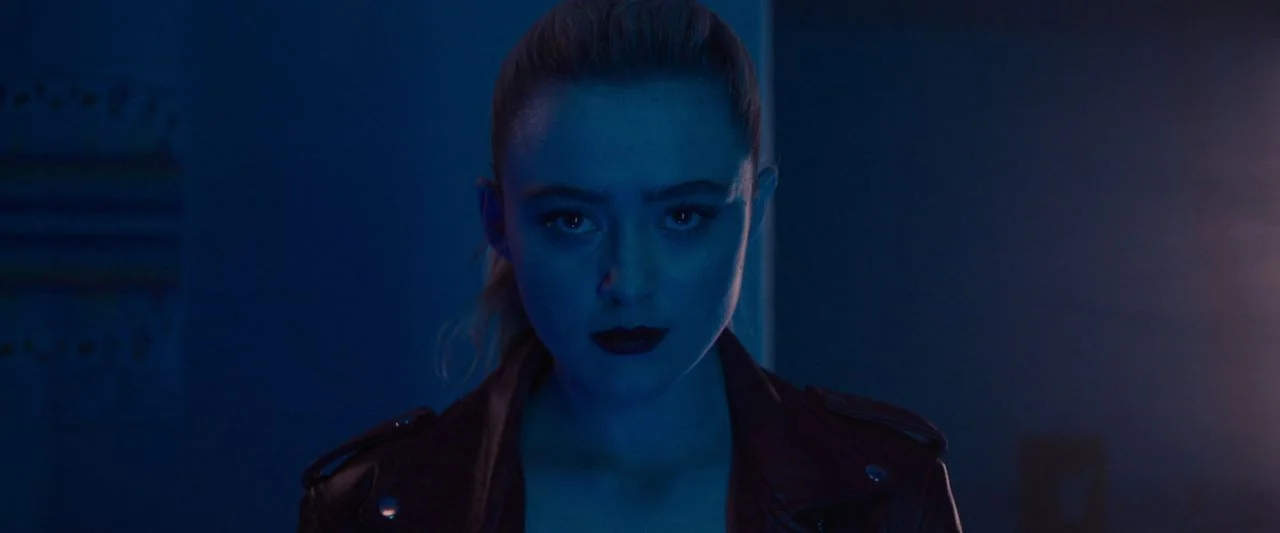 Freaky (2020) movie still: closeup — A dark indoor space illuminated by vibrant blue neon light. A young woman with blonde hair in a ponytail faces…