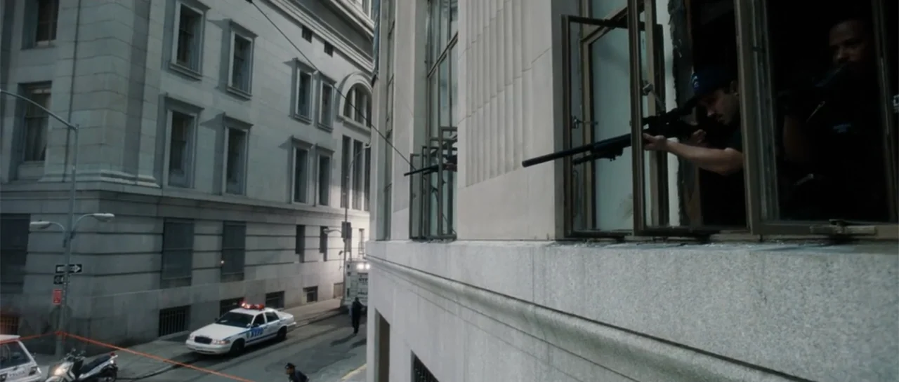 Inside Man (2006) movie still: wide — A stone building exterior with tall windows overlooks a city street lined with cars. A man leans out of a high…