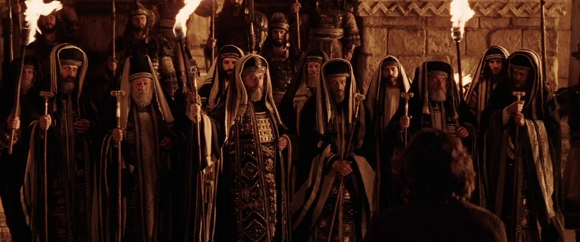 The Passion of the Christ (2004) movie still: wide — A group of older men in ornate robes stand in a stone-walled interior space. They hold long…