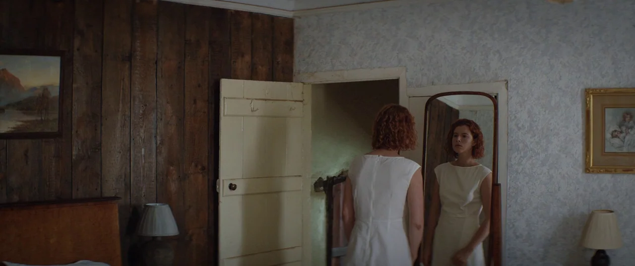 Beast (2017) movie still: medium — A bedroom features one wall with wood paneling and another with textured wallpaper. A woman wearing a white dress…
