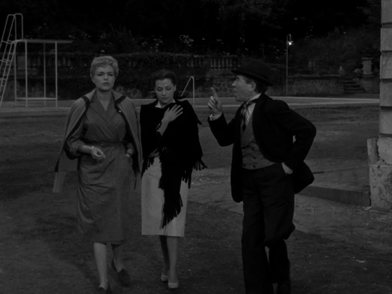 Diabolique (1955) movie still: fullbody — Three people walk across a paved outdoor space at night near a swimming pool area. The man on the right…
