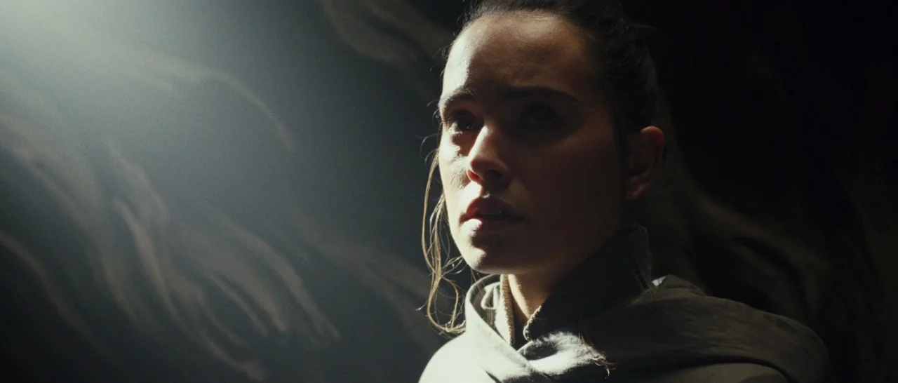 Star Wars: The Last Jedi (2017) movie still: closeup — A dimly lit cave space with dark rocky walls visible in the shadows. A woman gazes toward the…