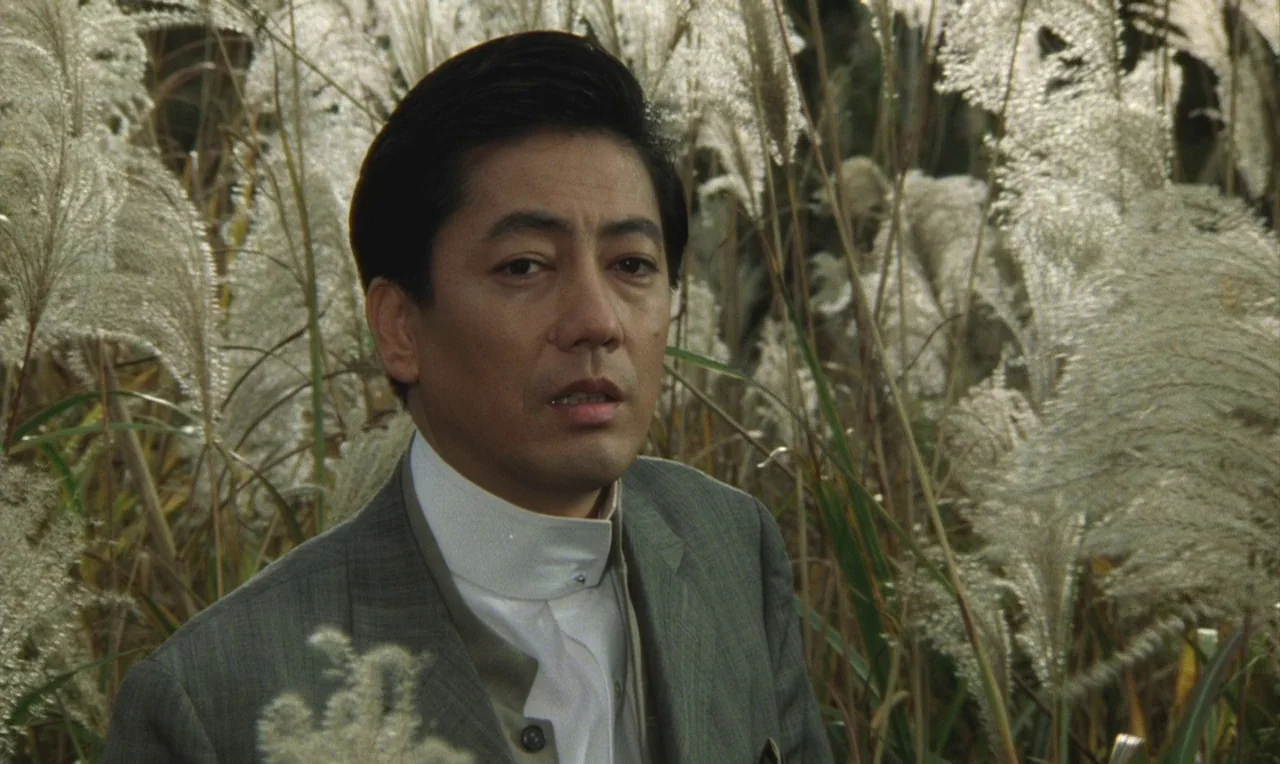 Yumeji (1991) movie still: medium — A man stands outdoors amidst a dense thicket of tall, silver-hued decorative grasses. He looks forward with an…