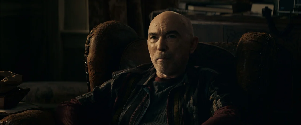 Hypnotic (2023) movie still: medium — A man sits inside a dark room in a damaged, vintage leather armchair. He stares ahead while resting in the…