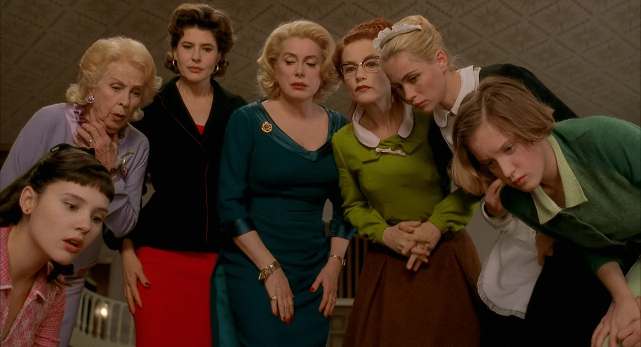 8 Women (2002) movie still: medium — Seven women are grouped together in a room with patterned wallpaper. They stand close together and peer…