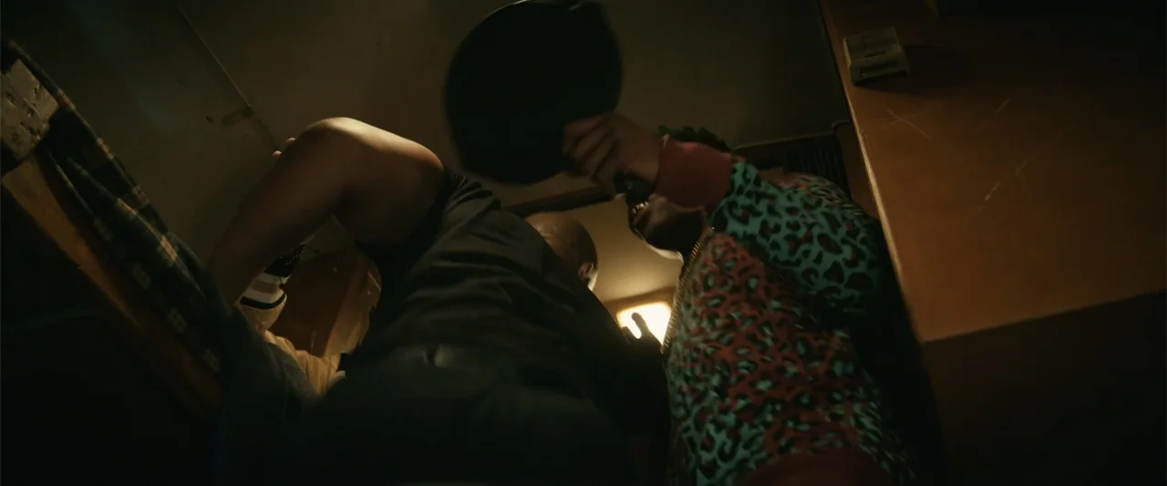Last Looks (2022) movie still: medium — A cramped interior space with dark walls and low overhead lighting. One man holds a frying pan above another…