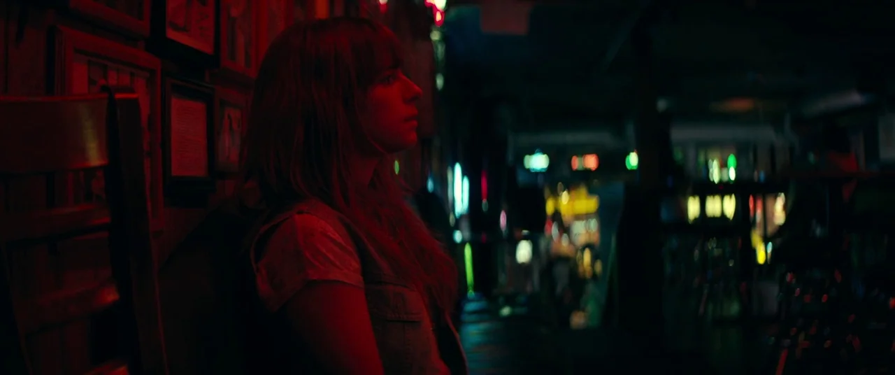 Wild Rose (2018) movie still: medium — A dimly lit establishment with framed pictures on a wall and blurred neon background. A woman sits in a dark…