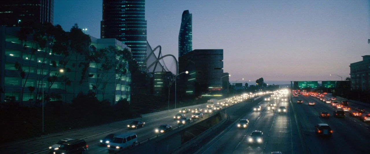 Southland Tales (2006) movie still: establishing — A wide view of a multi-lane highway at dusk near a modern city skyline. Multiple cars with bright…