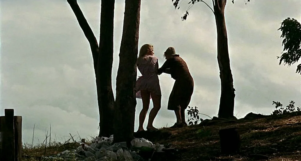 Age of Consent (1969) movie still: wide — A grassy hillside dotted with trees features a pile of scattered trash. Two women stand near the trees…