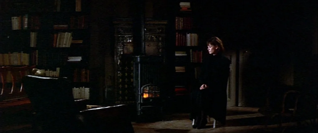Love Unto Death (1984) movie still: wide — A dimly lit room features wall-to-wall wooden bookshelves packed with books. A woman sits in a chair near…