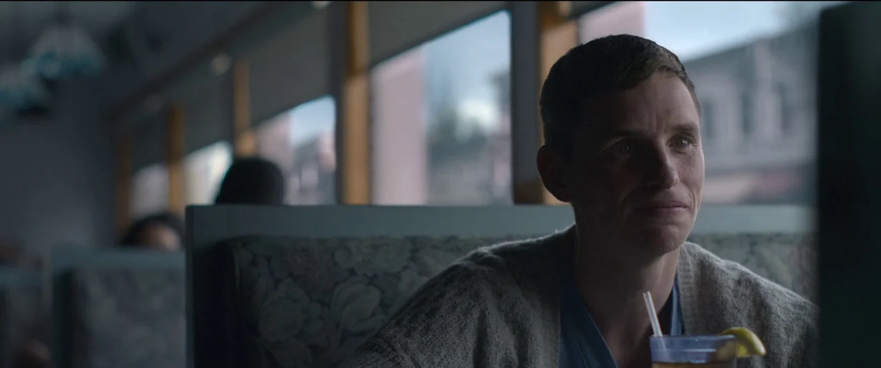 The Good Nurse (2022) movie still: A pensive young man sits in the corner of a quiet diner booth on an overcast day. The soft, diffused light from…