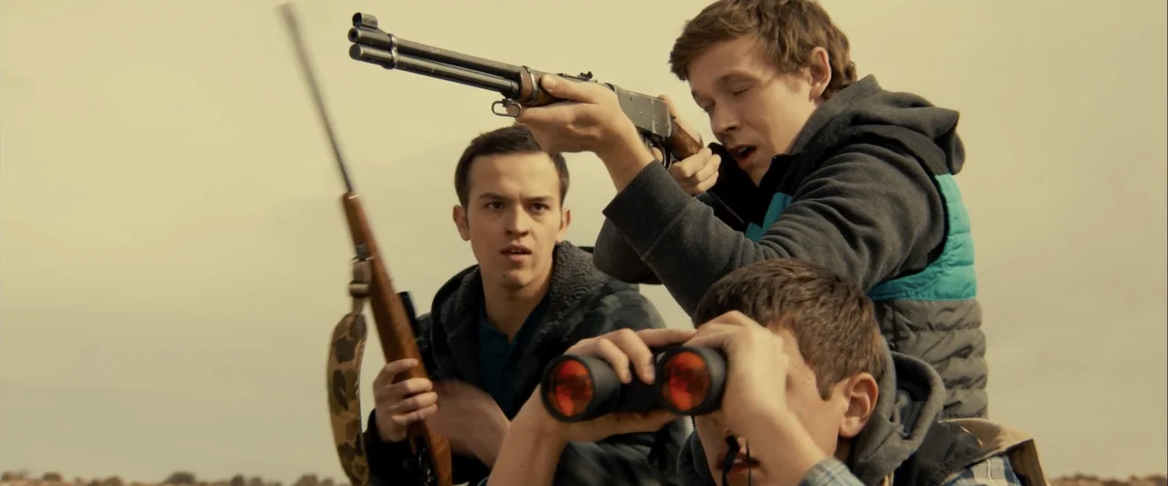 Frontera (2014) movie still: medium — An open field under an overcast sky provides the backdrop for three young men. The group prepares to hunt with…