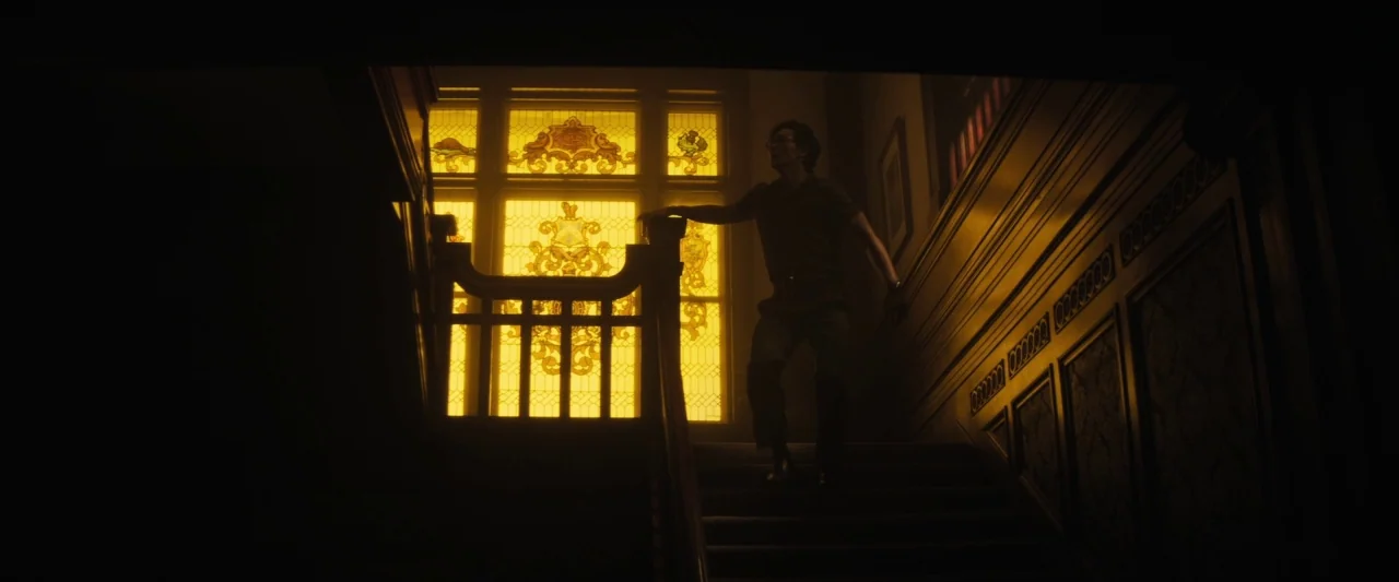 The Grudge (2020) movie still: medium — A dimly lit interior staircase featuring dark wood paneling and a large stained glass window. A man in dark…