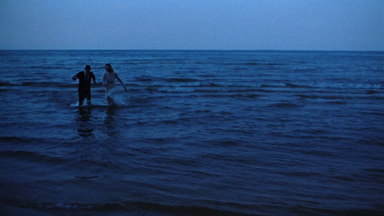 Strawberry Mansion (2021) movie still: wide — A vast expanse of dark blue water stretches to the horizon under twilight skies. A man in a suit and a…