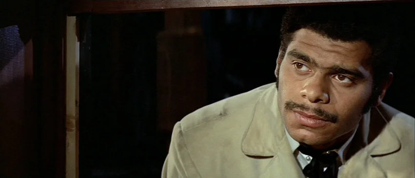 Whity (1971) movie still: closeup — A man stands partially obscured behind a dark vertical wooden structure. He looks toward the side of the frame…