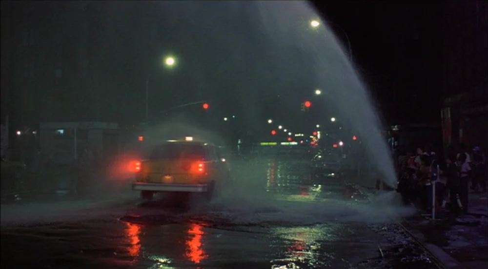 Taxi Driver (1976) movie still: wide — A dark urban street at night features a taxi driving through water spray. A group of people stands on the…