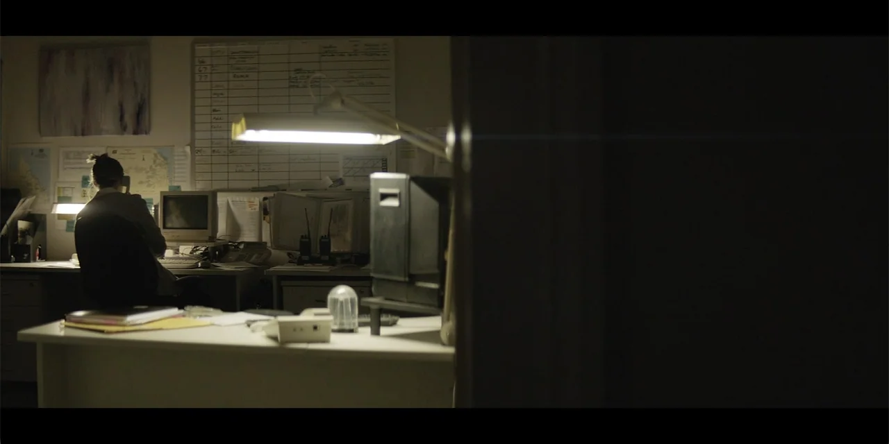 La coda del diavolo (2024) movie still: wide — A dark office setting with a desk occupied by a person working at night. The subject sits at a desk…