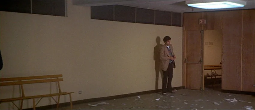 California Split (1974) movie still: wide — A sparsely furnished corridor features beige walls and an open doorway with wood paneling. A lone man…