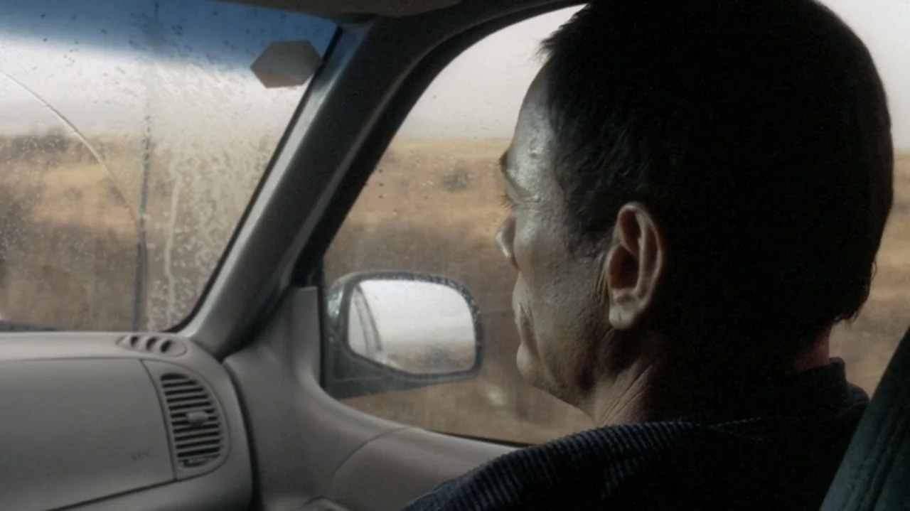 The Shepherd (2023) movie still: medium — The interior of a moving car shows a man looking out at a field. A dirty side window reveals a blurry…