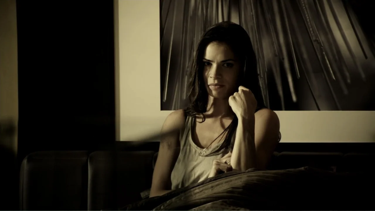 Assassination Games (2011) movie still: medium — A dimly lit room features a black sofa placed against a wall with framed art. A young woman with…