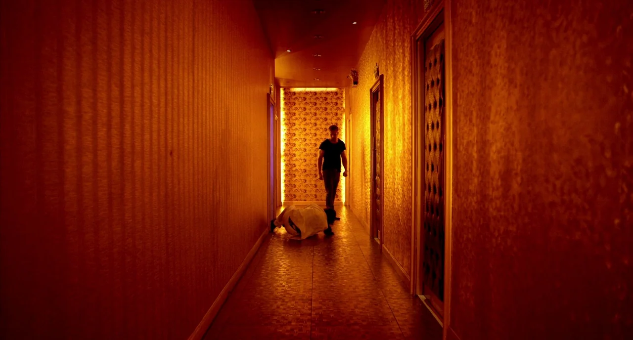 Only God Forgives (2013) movie still: wide — A narrow hotel corridor features textured wallpaper and bright illuminated floral wall accents. A man…
