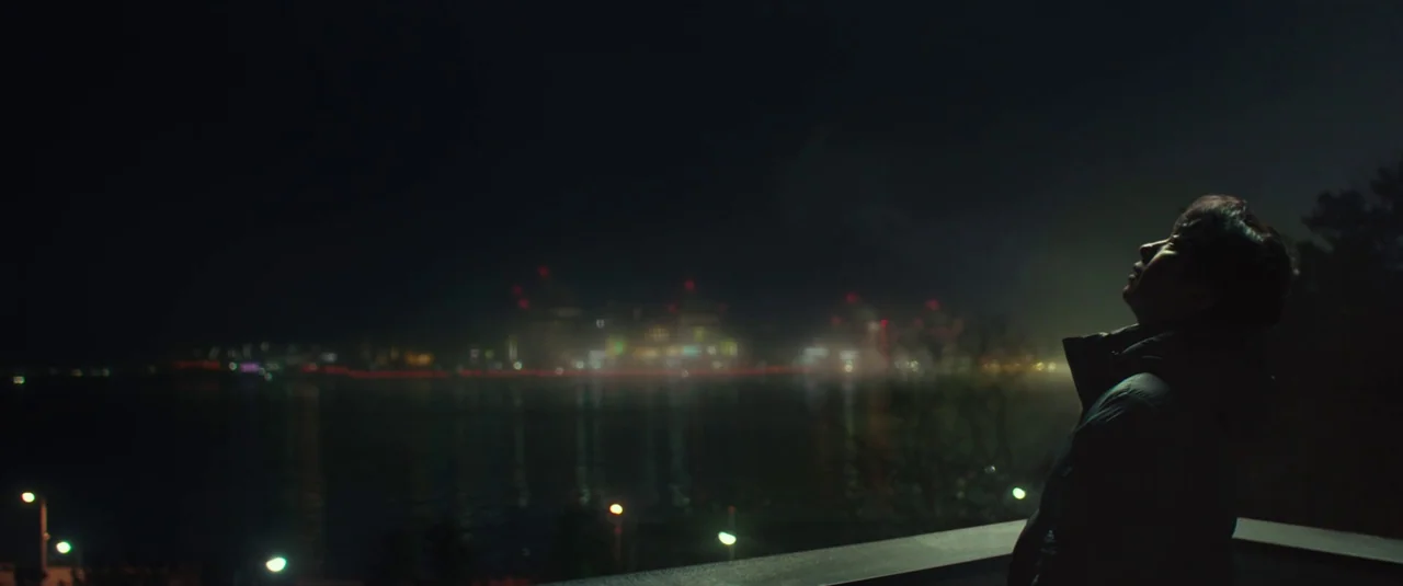 Decision To Leave (2022) movie still: medium — A dark outdoor platform overlooks a body of water at night. A young man stands at the railing gazing…