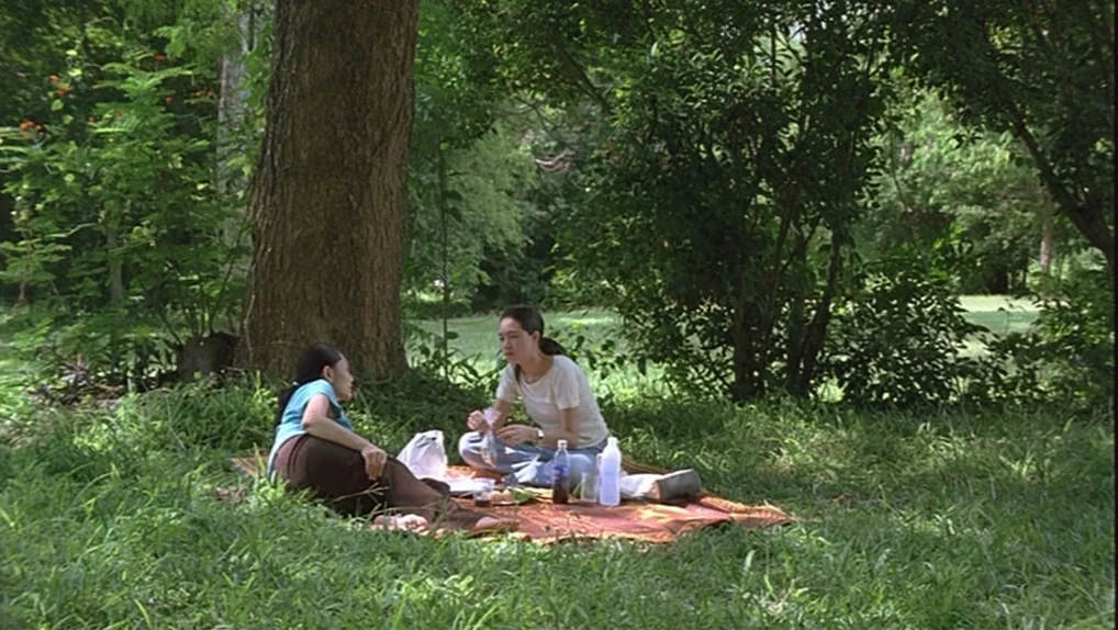 Syndromes and a Century (2006) movie still: wide — A grassy area beneath a large tree provides shade for a picnic setup. Two young women sit on a…