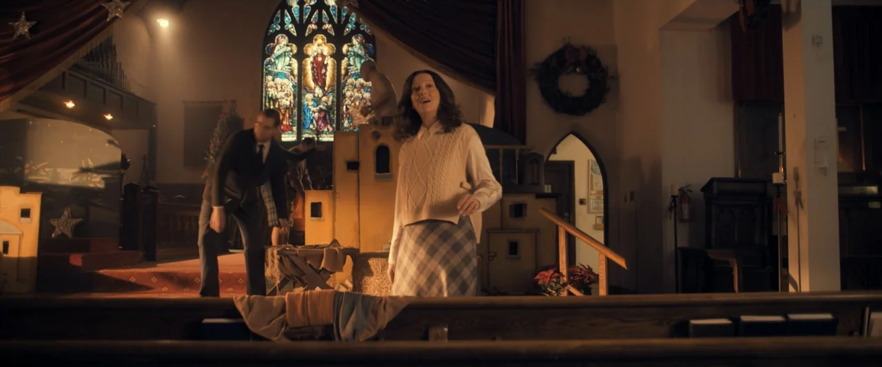 The Best Christmas Pageant Ever (2024) movie still: medium — A church interior features wooden pews in the foreground and a decorated stage area. A…