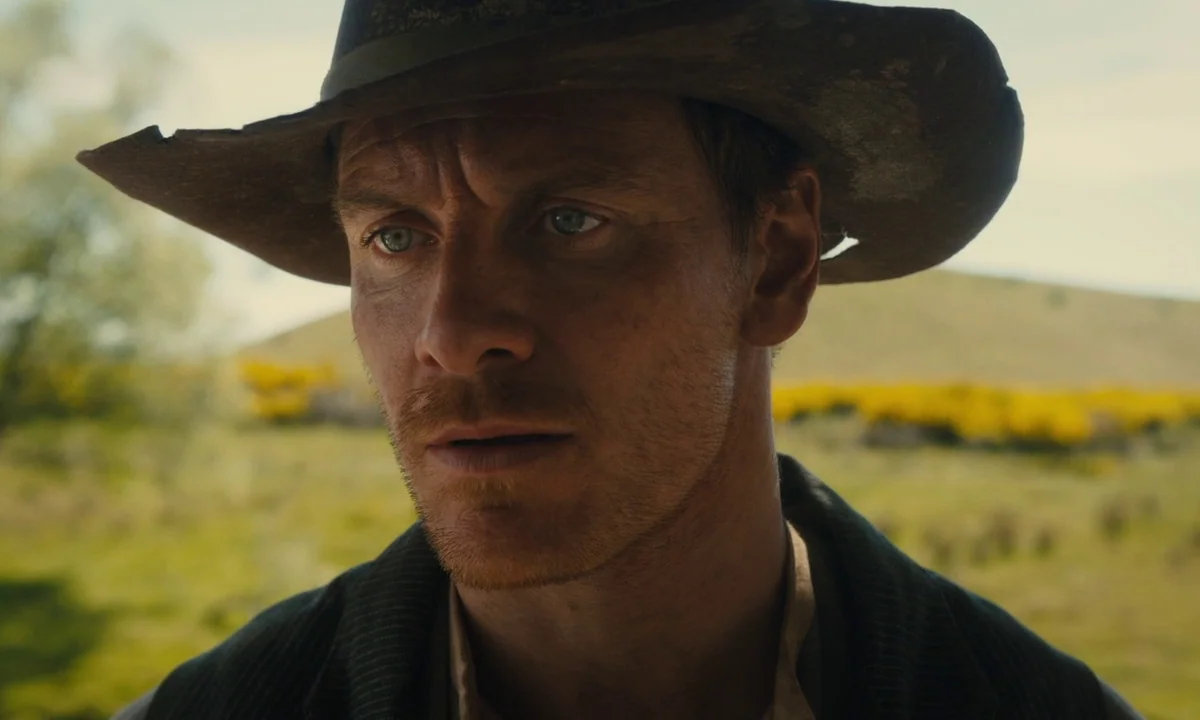 Slow West (2015) movie still: closeup — A man in a weathered hat stands outdoors in a bright, grassy landscape. The man gazes past the camera lens…