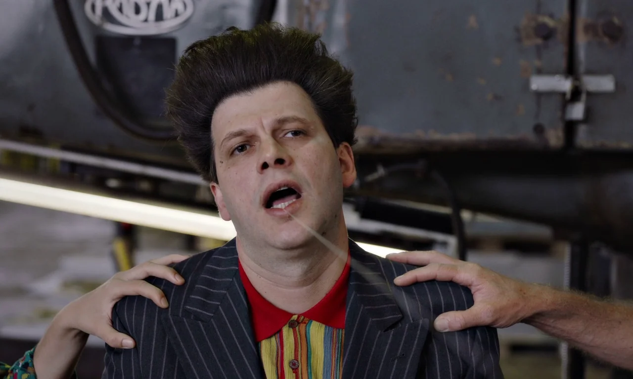 Bloodsuckers (2021) movie still: medium — The interior of an industrial auto repair shop shows a vehicle lift in the background. A man stands at the…
