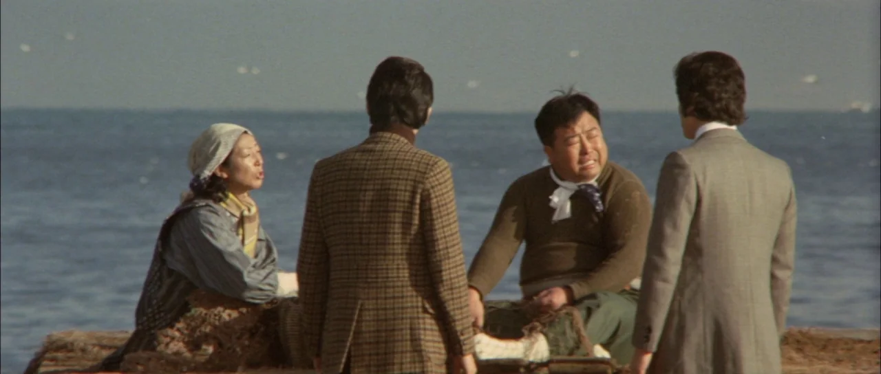 Terror of Mechagodzilla (1975) movie still: medium — A group of four people stands on a weathered wooden dock by the sea. The individuals interact…