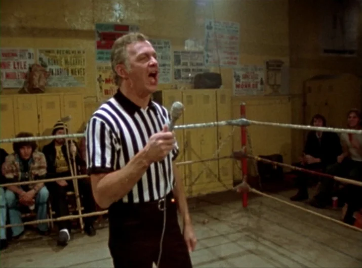 Desperate Living (1977) movie still: medium — A worn gym interior features metal lockers covered in aged boxing promotion posters. A referee stands…