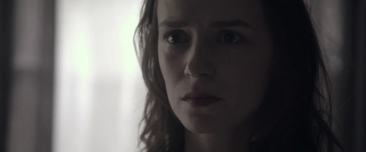 Starry Eyes (2014) movie still: closeup — A dimly lit room with a bright light source on the left side. A woman stands in the shadows looking…